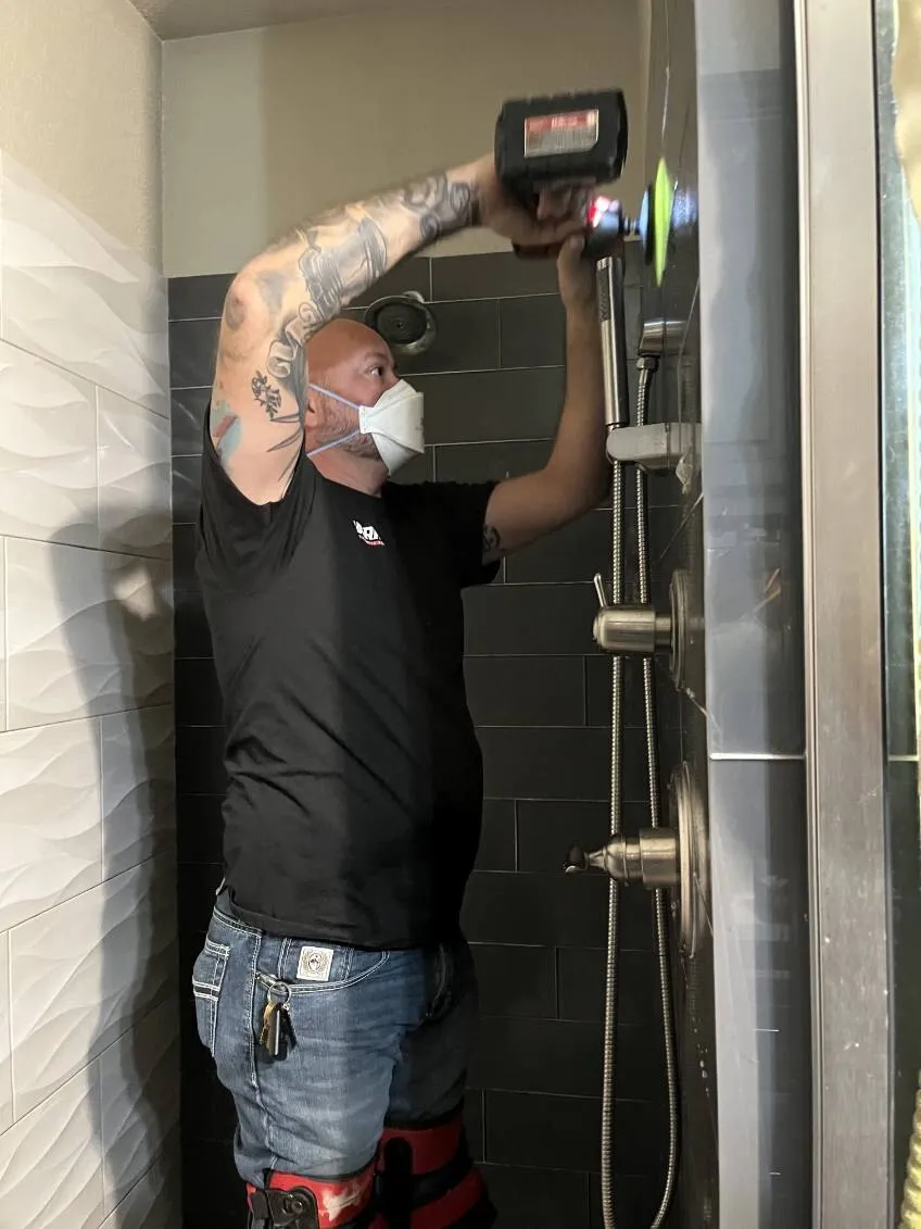 A technician in a respirator scanning a tiled shower wall with a thermal camera.