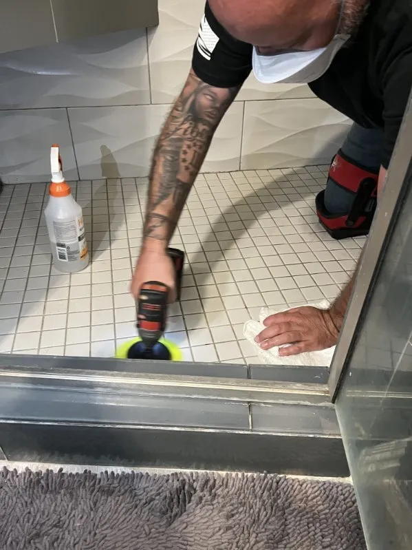 A technician running a cleaning tool along a tile floor at the base of a shower.