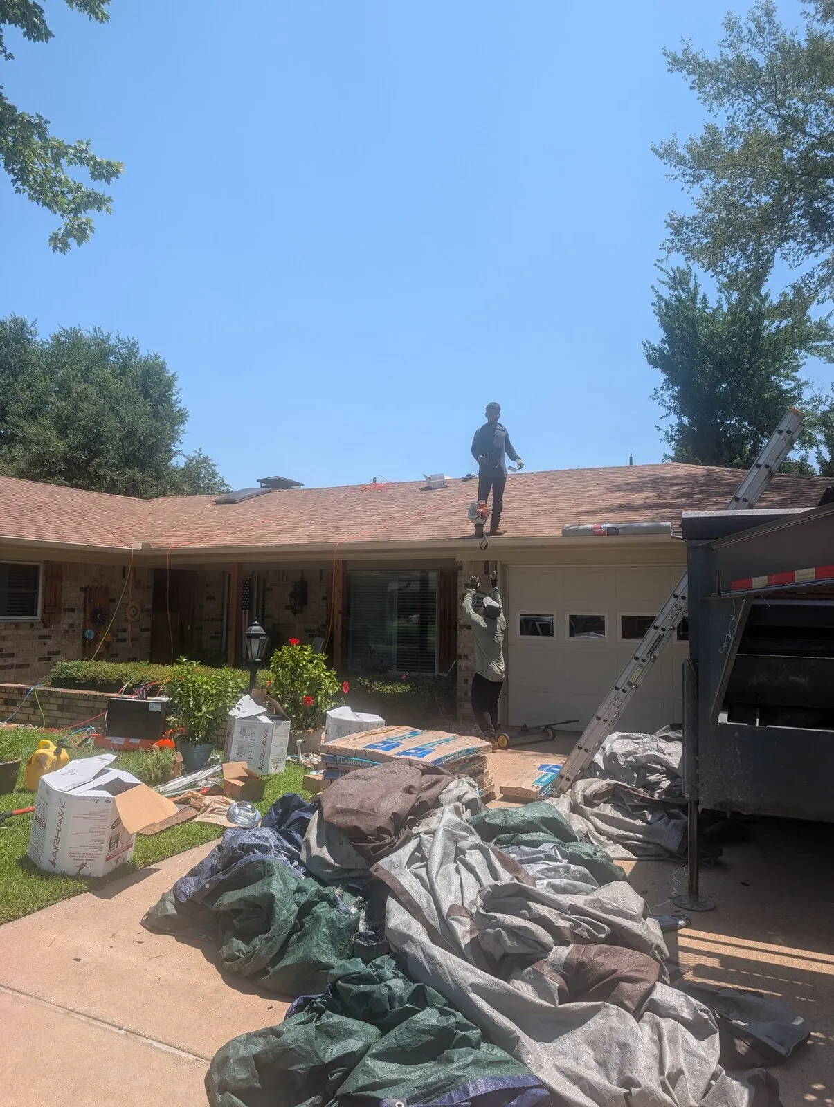 A roofer standing on the ridge of a house with torn-off shingles bagged in the driveway.