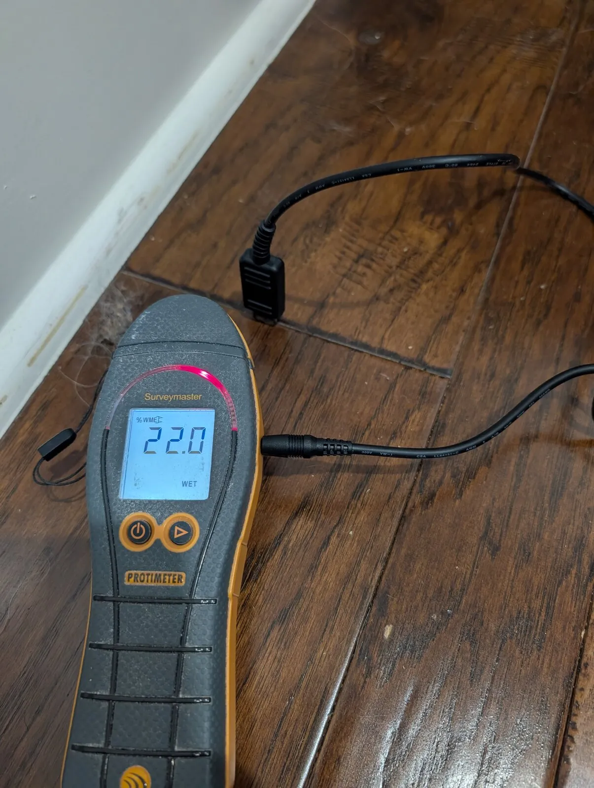 A moisture meter set on a hardwood floor beside the baseboard.