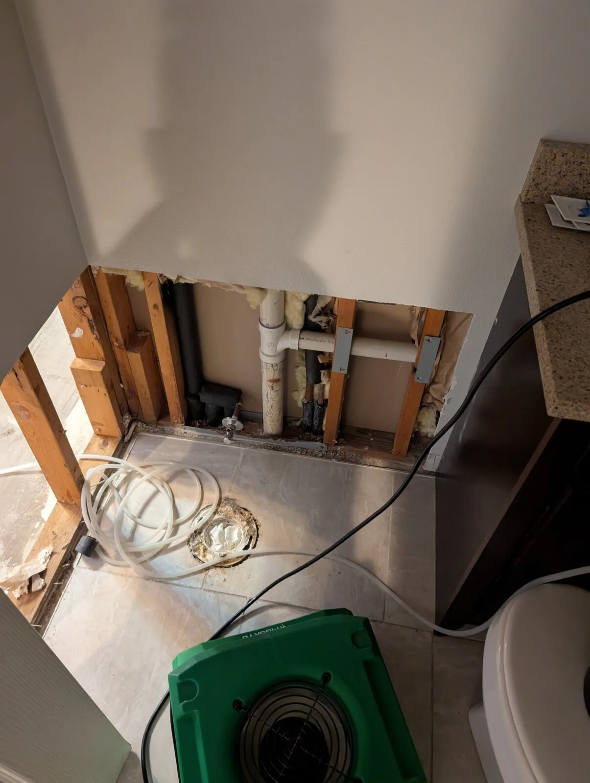 An opened wall under a sink with an air mover running on the floor below it.